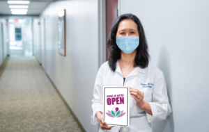 Dr. Cynthia wearing face mask and holding sign that says Come In We're Open