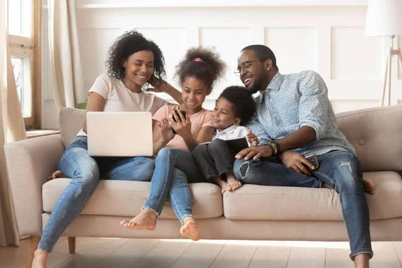 smiling family of four sitting together on couch all with phones or laptops