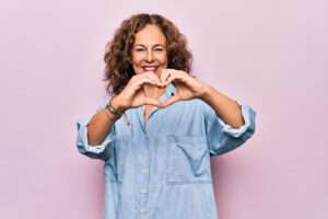 middle aged smiling woman in denim shirt holding her hands in shape of a heart